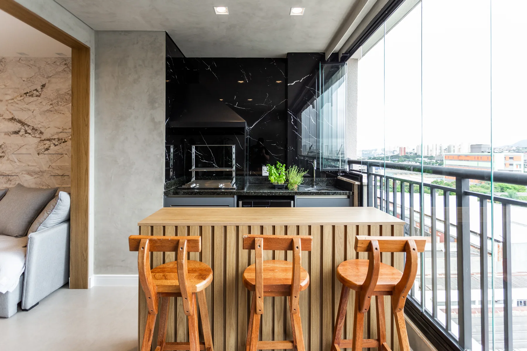Área gourmet planejada em varanda de apartamento com bancada ripada de madeira, banquetas, churrasqueira e vista da cidade, em Atibaia