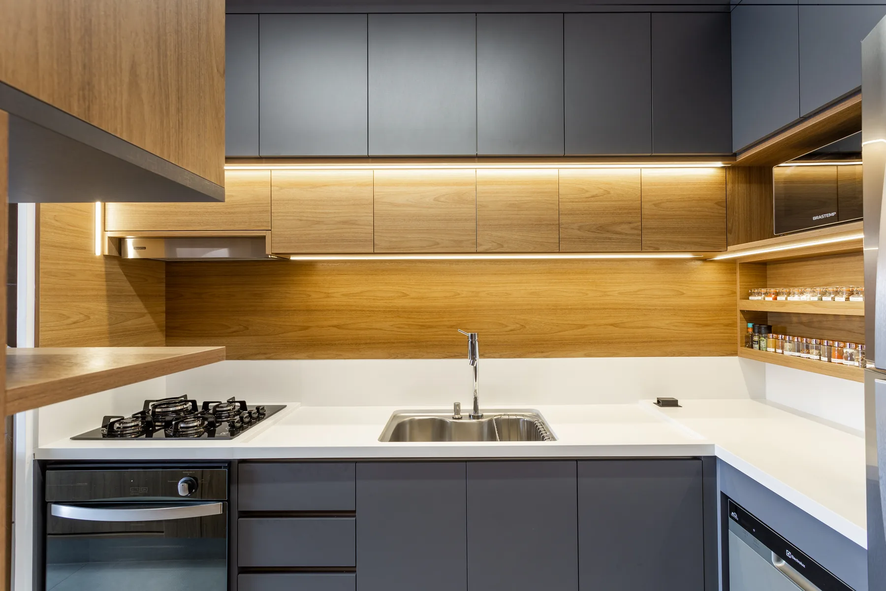 Cozinha planejada sob medida em L, com marcenaria cinza e madeira, nicho de temperos e iluminação de LED sob os armários, em Atibaia